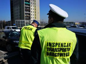 przy zaparkowanym samochodzie stoi policjant i funkcjonariusz Inspekcji Transportu Drogowego, na sobie mają odblaskowe kamizelki, policjant schyla się do kierowcy kontrolowanego pojazdu i z nim rozmawia