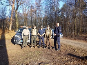 w lesie pozują do zdjęcia dwaj umundurowani policjanci i trzech strażników leśnych, za mundurowymi stoją zaparkowane pojazdy Policji i Straży Leśnej