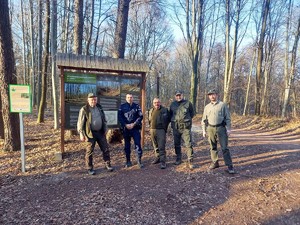 na tle leśnej tablicy informacyjnej pozują do zdjęcia umundurowany policjant i czterech strażników leśnych