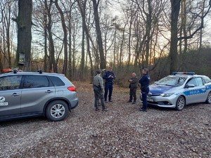 w lesie stoją dwa pojazdy, radiowóz policyjny i pojazd Straży Leśnej, pomiędzy nimi stoją dwaj umundurowani policjanci i trzech strażników leśnych, rozmawiają ze sobą