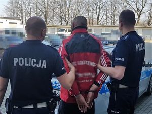 na parkingu policyjnym dwóch umundurowanych policjantów trzyma za ramiona mężczyznę, który stoi tyłem i na rekach ma założone kajdanki