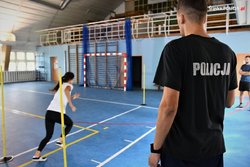 na sali gimnastycznej stoją tyczki, kobieta w stroju sportowych rozpoczyna bieg, z boku stoi instruktor w koszulce z napisem Policja