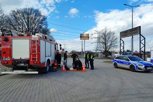 na parkingu samochodowym stoi wóz strażacki oraz policyjny radiowóz, między samochodami stoją strażacy z OSP, policjanci, przed nimi leży koc, na którym leży fantom, mężczyzna kuca przy fantomie, naprzeciwko niego jest strażak, który udziela mu instrukcji