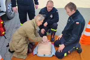 Kobieta układa ręce na klatce piersiowej fantoma, strażacy przyglądają się, klęcząc na przeciwko niej, z drugiej strony fantoma