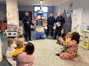 w sali przedszkolnej na podłodze siedzą dzieci, jedno z dzieci siedzi na wózku inwalidzkim. Przed dziećmi stoją trzej umundurowani policjanci oraz maskotka śląskiej Policji Pies Sznupek