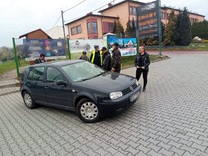 na parkingu samochodowym stoi zaparkowane auto, umundurowany policjant stoi obok osób ubranych w ubiór motocyklowy, którzy rozmawiają z kierowcą samochodu