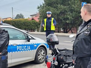 na parkingu samochodowym stoi zaparkowany radiowóz, obok stoją motocykle. Przy drodze stoi umundurowany policjant, pokazuje kciuki uniesione w górę, przed nim stoją osoby w ubiorze motocyklowym