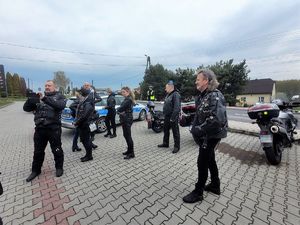 na parkingu samochodowym stoją osoby w ubiorze motocyklowym, zaparkowane motocykle, w oddali stoi umundurowany policjant, który obserwuje ruch pojazdów