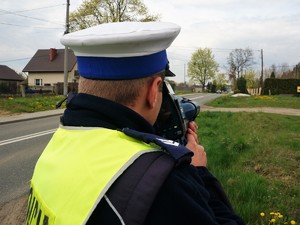 umundurowany policjant mierzy prędkość urządzeniem do pomiaru prędkości, stoi przy jezdni