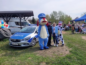 podczas festynu w plenerze do wspólnego zdjęcia pozują Sznupek, umundurowany dzielnicowy i osoba przebrana w strój krówki, obok stoi zaparkowany radiowóz, do elementów ogrodzenia są przyczepione kolorowe balony