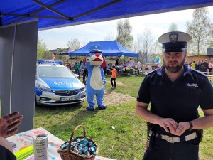 umundurowany policjant stoi przy stoisku profilaktycznym, w tle macha łapką Sznupek - maskotka Śląskiej Policji, obok stoi zaparkowany radiowóz