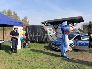 w policyjnym radiowozie siedzi dziecko, obok radiowozu stoi kobieta, z drugiej strony, blisko stoiska profilaktycznego Policji stoi umundurowany policjant i trzyma puszkę na datki w rękach, obok radiowozu stoi też pies Sznupek, maskotka Śląskiej Policji