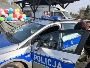 chłopiec siedzi w radiowozie na miejscu kierowcy, mężczyzna stoi obok radiowozu i patrzy na chłopca, w tle widać kolorowe balony przyczepione do ogrodzenia