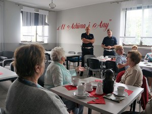 policjanci stoją przed seniorami siedzącymi przy stołach, na których stoją filiżanki