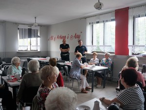 policjanci stoją przed seniorami siedzącymi przy stolikach, prowadzą spotkanie profilaktyczne