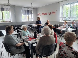 policjantka stoi przed seniorami siedzącymi przy stolikach, prowadzi spotkanie profilaktyczne