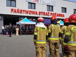 na placu przed komendą straży pożarnej w rzędzie stoją strażacy w strojach ćwiczebnych, na przeciwko stoją strażacy w mundurach galowych