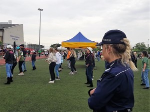 na pierwszym planie stoi policjantka, która przygląda się grupie osób, które są ustawione w szyku i wykonują taniec