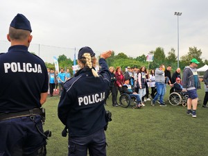 po boisku maszerują uczestnicy XVIII Powiatowej Olimpiady Osób Niepełnosprawnych, na pierwszym planie, tyłem do zdjęcia stoją  policjant i policjantka, która macha ręką do uczestników