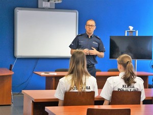 policjant prowadzi spotkanie z młodzieżą, uczennice w koszulkach z napisem klasa policyjna siedzą w ławce i go słuchają