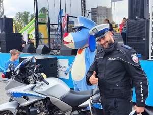 policjant w mundurze uśmiecha się i pokazuje kciuk w górę, obok stoi policyjny motocykl i maskotka śląskiej Policji, pies Sznupek