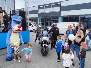 maskotka śląskiej Policji, pies Sznupek trzyma za ręce małe dziecko, inne dziecko odchodzi od Sznupka, uśmiecha się, obok stoi policjant w mundurze motocyklowym, dorośli i dzieci