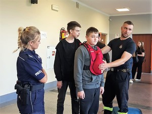 policjantka stoi obok chłopaka, która ma na sobie sprzęt służący do doskonalenia umiejętności udzielania pierwszej pomocy podczas zadławienia, za chłopakiem stoi drugi chłopak, obok nich stoi strażak