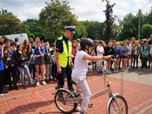 policjant asekuruje dziewczynę jadącą na rowerze, obok stoi grupa młodzieży i się im przygląda