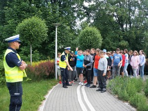 w miasteczku rowerowym policjant stoi obok grupy młodzieży i opowiada im o znakach drogowych, obok nich stoi drugi policjant, który im się przygląda