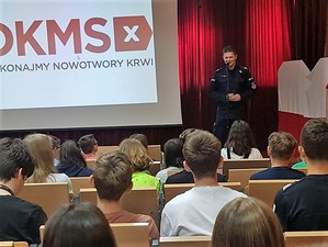 umundurowany policjant stoi na scenie, za sobą ma wyświetlone logo DKMS, przed nim, na widowni siedzi młodzież