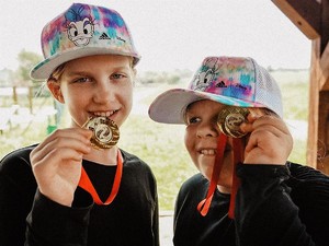 dwie dziewczynki trzymają przy twarzy złote medale, na głowach mają czapki z daszkiem, uśmiechają się