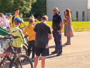 na szkolnym boisku stoi umundurowany policjant, przed nim stoją dzieci, które trzymają swoje rowery, za policjantem stoi kobieta