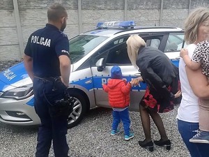 policjant stoi obok policyjnego radiowozu, przed nim kuca kobieta, która znajduje się za małym dzieckiem, i opowiada mu na ucho o pojeździe
