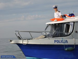 na zbiorniku wodnym znajduje się policyjna motorówka, na rufie stoi policjantka w mundurze przeznaczonym do pełnienia służby na łodzi motorowej, tj. biała koszulka i czarne spodenki.