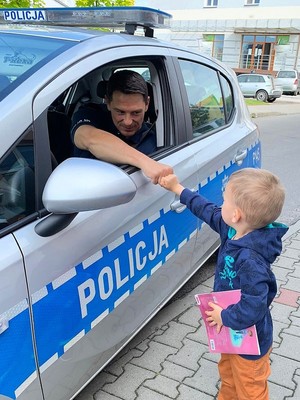 Policjant siedzi w radiowozie, obok radiowozu stoi chłopczyk, którzy przebija żółwika z policjantem.