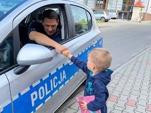 Policjant siedzi w radiowozie, obok radiowozu stoi chłopczyk, którzy przebija żółwika z policjantem.