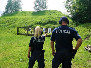 policjantka w prawej dłoni trzyma broń, celuje do tarczy, którą ma przed sobą, obok policjantki stoi policjant, przygląda się jej strzelaniu