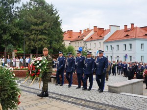 pięciu policjantów w mundurach galowych podchodzą do Pomnika Powstańców Śląskich, przed nimi idzie żołnierz, który niesie wieniec z kwiatami w kolorach białych i czerowonych