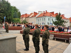 trzech żołnierzy stoi w szyku, oddają honor, z tyłu stoją zebrani goście podczas uroczystych obchodów