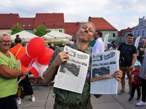 młody mężczyzna w mundurze harcerskim pozuje do zdjęcia z gazetami przygotowanymi na obchody 100-lecia przyłączenia Górnego Śląska do Polski, w tle stoją zebrani uczestnicy tego wydarzenia