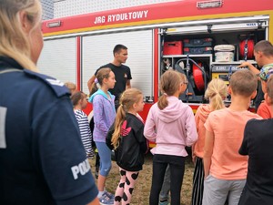 grupa dzieci stoi przy wozie strażackim, słuchają opowiadań strażaków, na pierwszym planie stoi policjantka, która również słucha strażaków