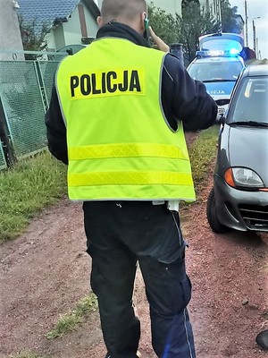 policjant w odblaskowej kamizelce stoi tyłem, rozmawia przez telefon komórkowy, w tle widać policyjny radiowóz oraz kawałek samochodu osobowego