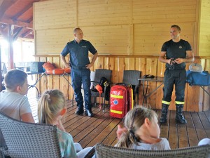 policjant oraz strażak stoją przed siedzącymi na krzesłach, przodem do nich dziećmi
