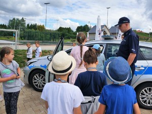 policjant stoi obok radiowozu, opowiada stojącym przy radiowozie dzieciom o swojej pracy