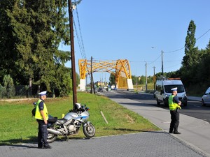 policjant obserwuje ruch pojazdów, obok niego stoi policyjny motocykl, obok motocykla stoi drugi policjant