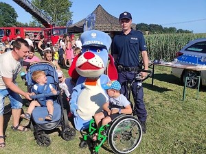 policjant i Sznupek pozują do zdjęcia z dziećmi, za nimi widać strażackie wozy i osoby podczas festynu charytatywnego