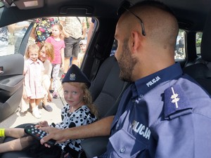 w policyjnym radiowozie, obok policjanta siedzi dziewczynka, na głowie ma założoną policyjną czapkę, obok radiowozu stoją inne dzieci, oglądają radiowóz