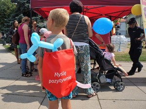 chłopiec stoi tyłem, na plecach ma założony plecak z napisem &quot;myśl pozytywnie&quot;, za nim stoją osoby przy stoisku profilaktycznym podczas festynu, stoi też policjantka