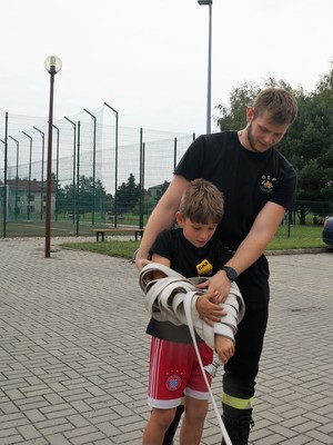 strażak pomaga chłopcu zwijać strażackiego wężą
