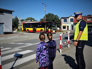 przed oznakowanym przejściem dla pieszych stoją dwie dziewczynki, obok nich stoi policjant, przygląda im się, widać również nadjeżdżający autobus miejski